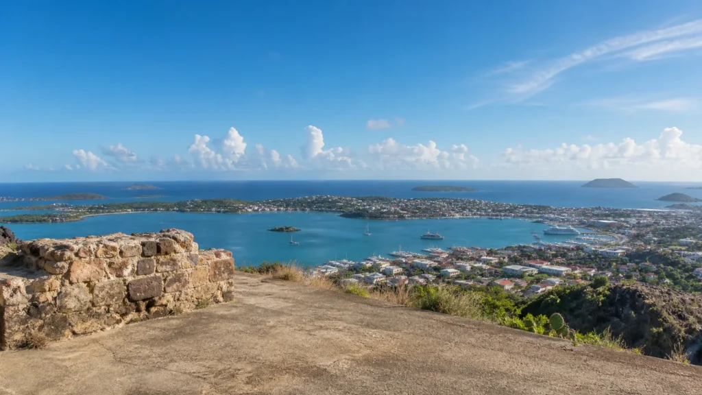 AI-generated editorial visualization of a non-facsimile weathered-masonry overlook above Marigot Bay; not an exact Fort Louis structure, current-condition, restoration, route, access or documentary record.